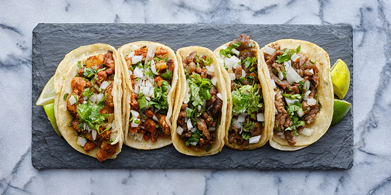 Plato con varios tacos de carne, tomate, cebolla morada, queso y un bol de salsa agria, entre otros ingredientes.